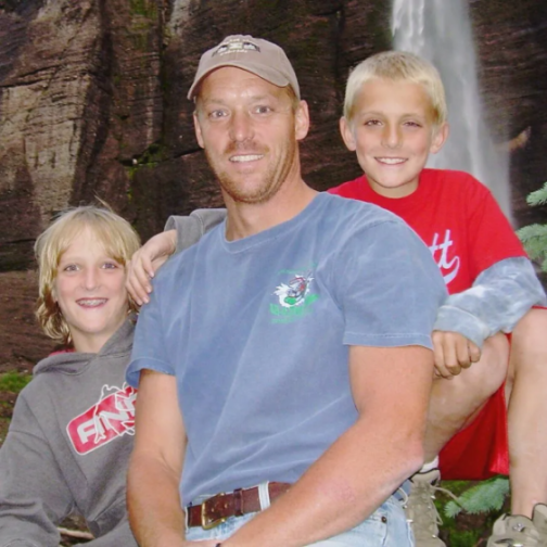 A man wearing a blue shirt and two young boys smiling with a waterfall in the background.