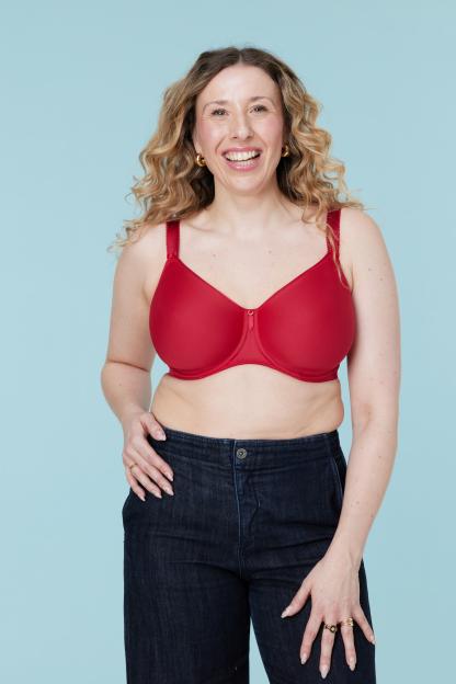 A smiling woman with curly blonde hair wears a red bra and dark jeans.