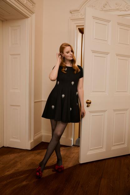 A woman with auburn hair in a black dress with jeweled floral embellishments, black tights, and red bow-tied shoes leans against a white doorframe.