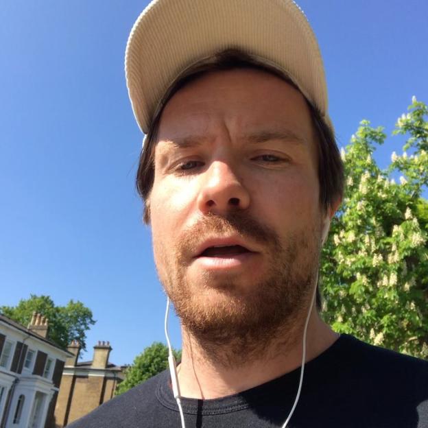 A close-up selfie of Joe Dempsie wearing a cap and white earbuds, with a clear blue sky and trees in the background.