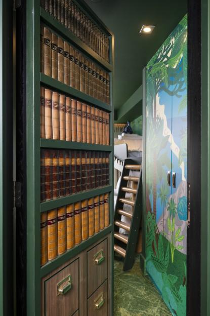 A dark green bookcase full of old leather-bound books that opens to reveal a children's bedroom with jungle-themed closet doors, a bunk bed, and a ladder.