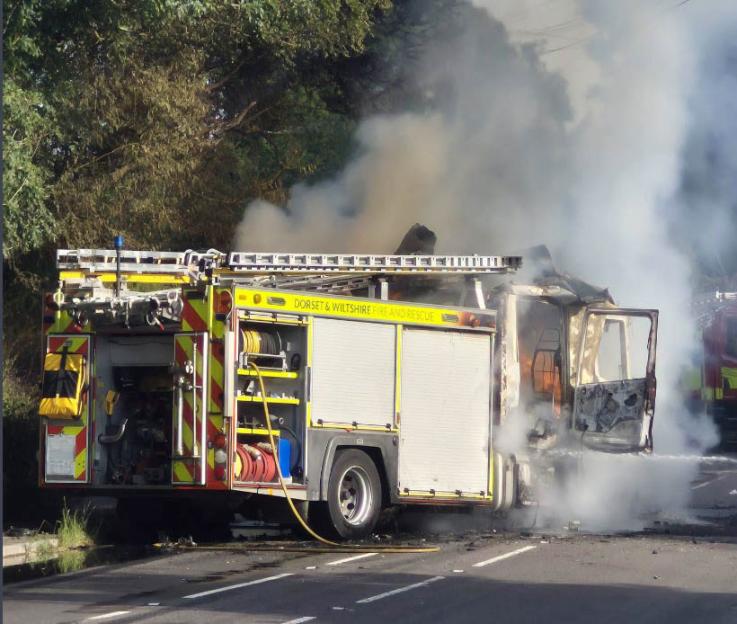 A fire engine from Dorset & Wiltshire Fire and Rescue, with its cab destroyed and emitting smoke, on a road.