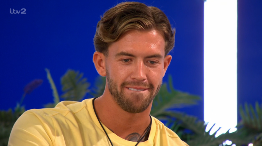 A man with wavy brown hair and a beard, wearing a yellow t-shirt, smiles faintly.