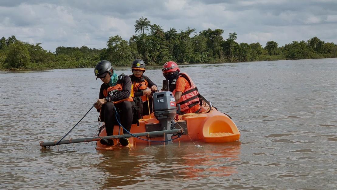 Three people in a small orange boat search a muddy river.