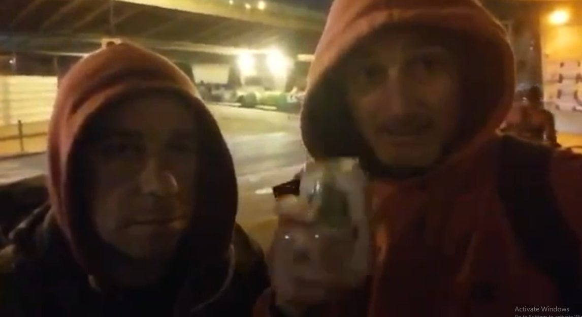 Two men in hoodies under an overpass at night, one holding a drink.