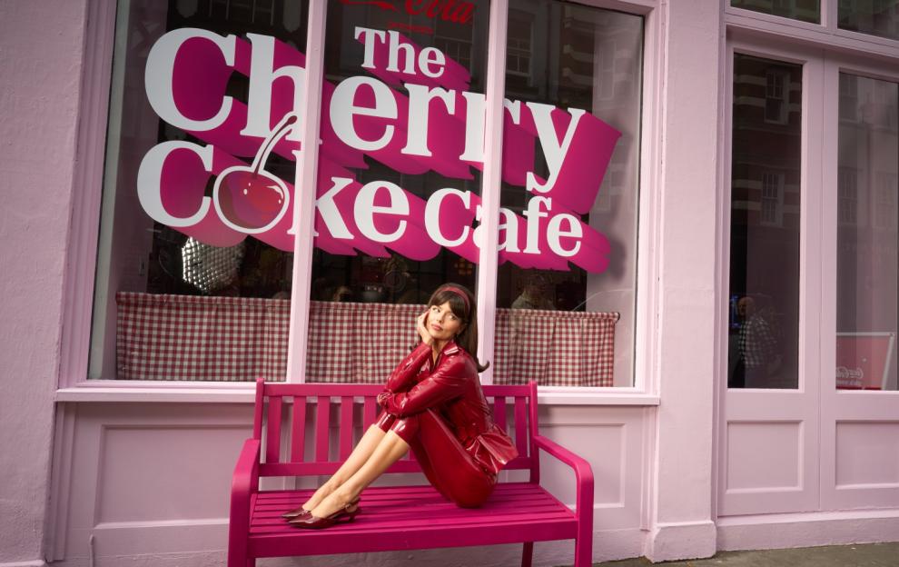 A woman in a red shiny outfit sitting on a pink bench outside "The Cherry Coke Cafe".
