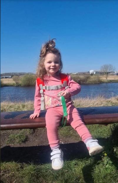 Isabelle Rose Welsh, a young girl with curly hair and a pink outfit, sitting on a bench with a body of water behind her.