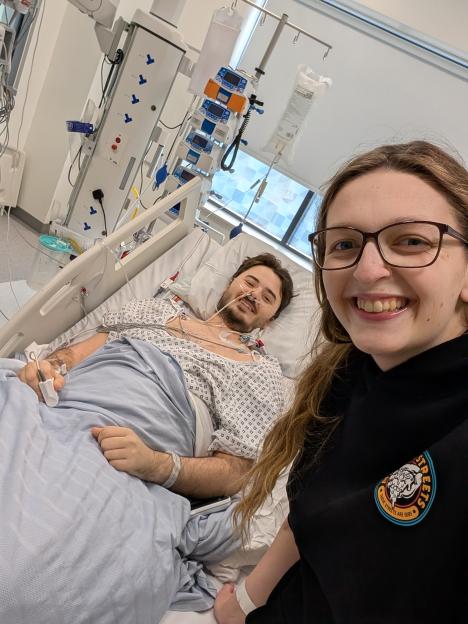 A man in a hospital gown lies in a hospital bed with medical tubes and wires attached to him, smiling while a woman in a black shirt takes a selfie beside him.