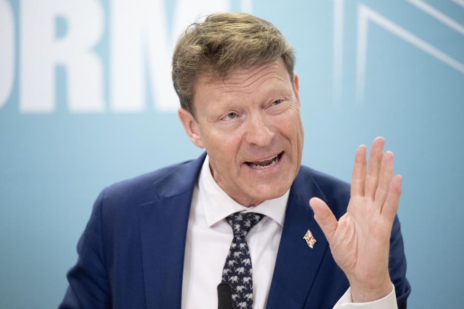 A man in a suit speaking and raising his hand, with a pin of the Union Jack flag on his lapel.