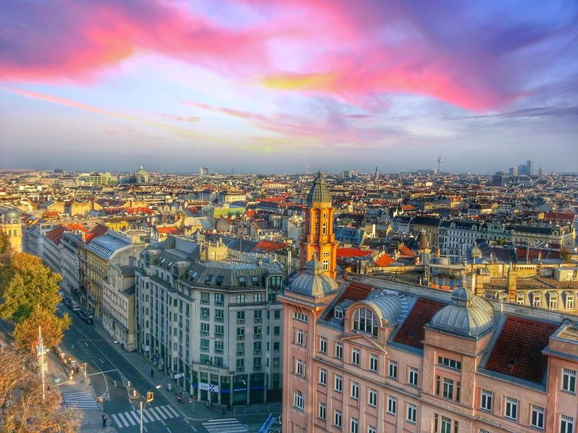 A high-angle view of the northern part of Vienna, Austria's skyline at sunset, with a mix of historic and modern buildings under a sky filled with pink, purple, and yellow clouds.