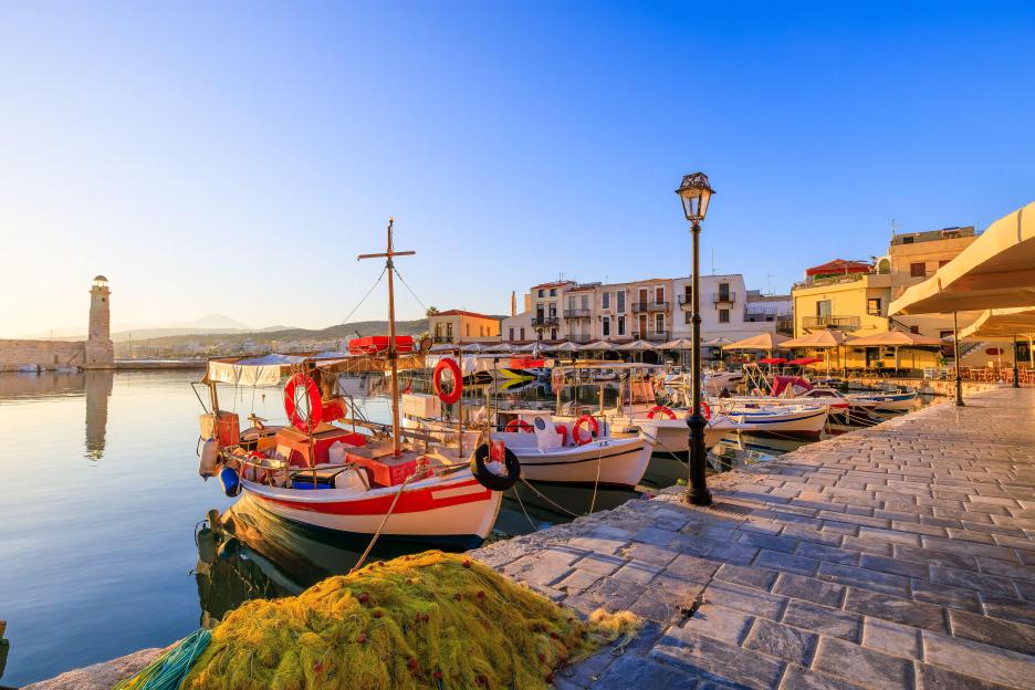 View of the old harbor in Rethymno, Crete, Greece, with boats docked and a lighthouse in the background.