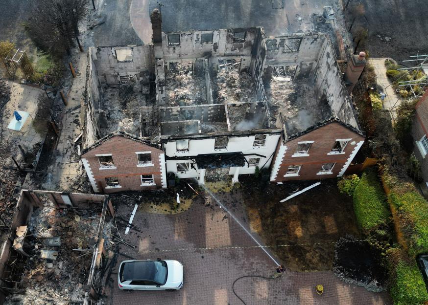 Aerial view of homes destroyed by wildfires, with a firefighter hosing down smoldering debris in the foreground.