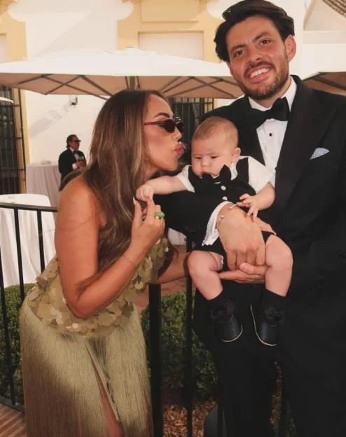 Pete Wicks wearing a tuxedo holding a baby while a woman in a gold dress leans in to kiss the baby's cheek.