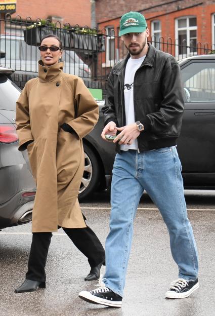 Maya Jama in a camel coat and Ruben Dias in a black leather jacket walk past a black car.