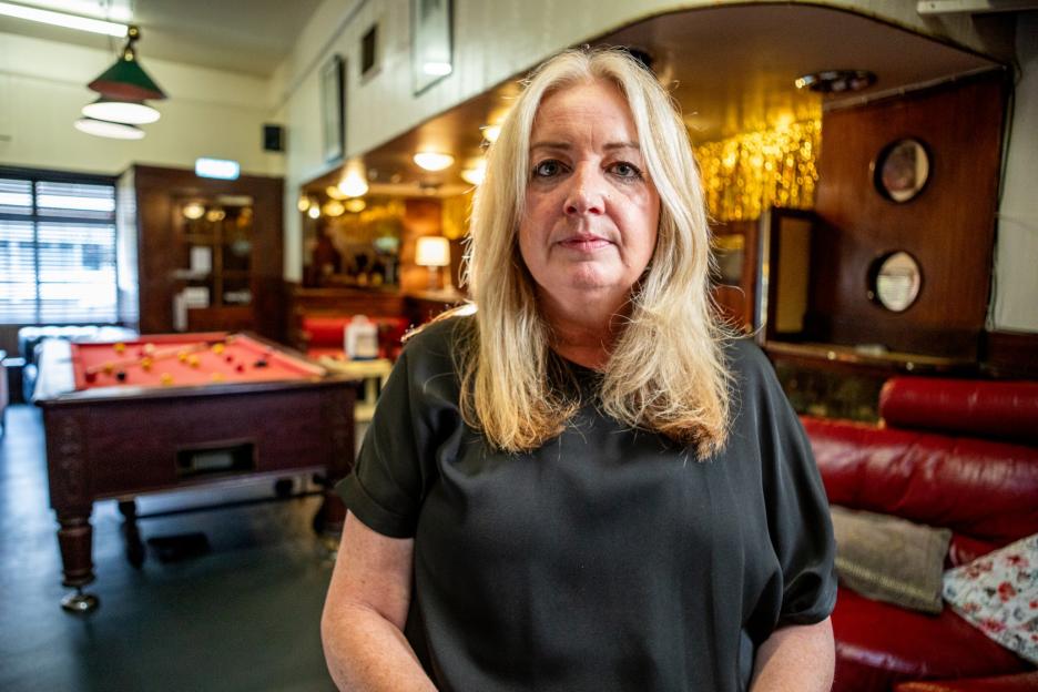 A middle-aged woman with blonde hair wearing a black top stands in a pub with a pool table and red leather couches.