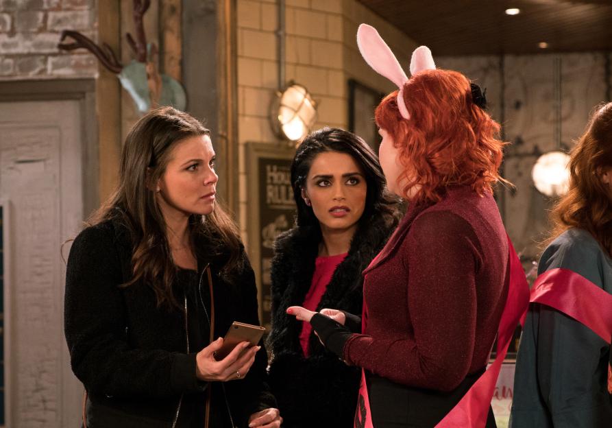 Three women in an empty factory, two of whom are wearing pink sashes, one with rabbit ears, and another holding a phone.