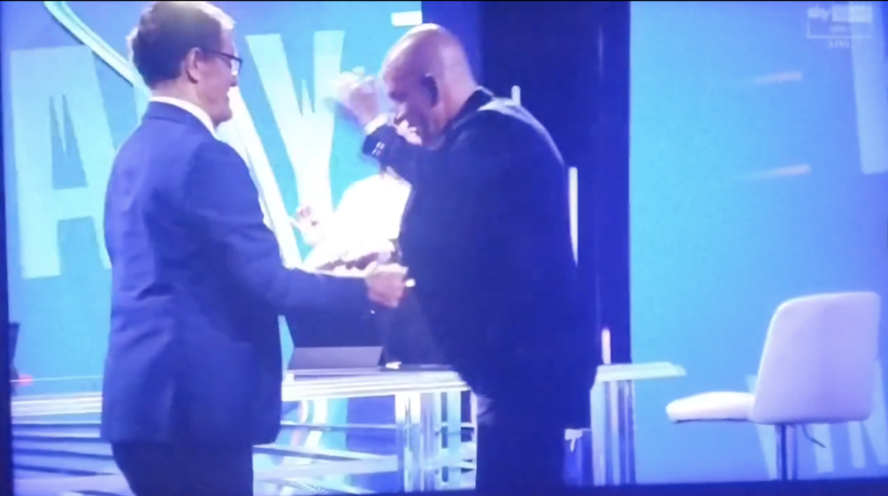 Two men, one in glasses and a suit, and a bald man in a dark shirt, stand facing each other and shaking hands on a stage with a blue backdrop.