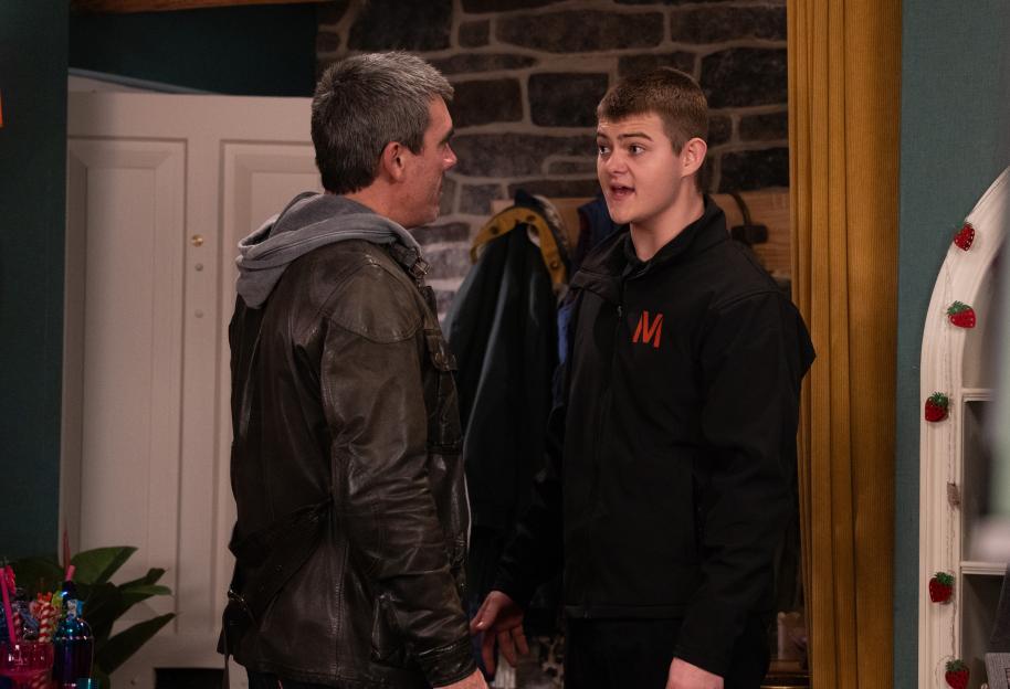 Two men talking, one in a leather jacket and the other in a black jacket with a red 'M' logo.