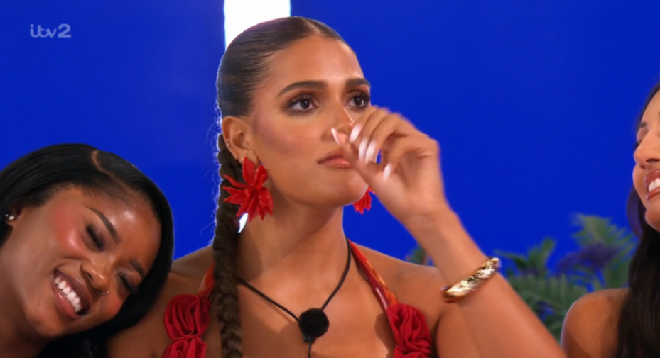 A woman in a red top wiping a tear, while another woman rests her head on her shoulder and smiles.