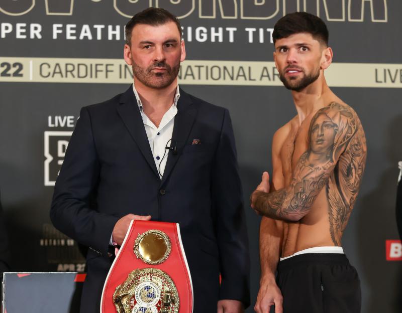 Boxer Joe Cordina, shirtless with a tattooed arm, stands beside a man in a suit holding a championship belt at a weigh-in event.