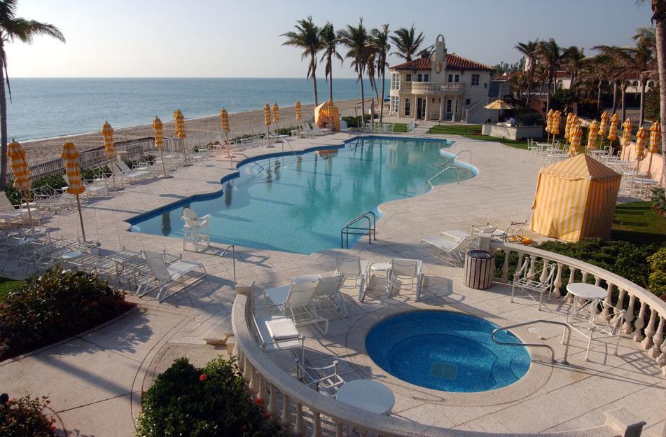 The Mar-a-Lago estate in Palm Beach, Florida, featuring a large swimming pool, spa, and patio with lounge chairs and umbrellas, all overlooking the ocean.