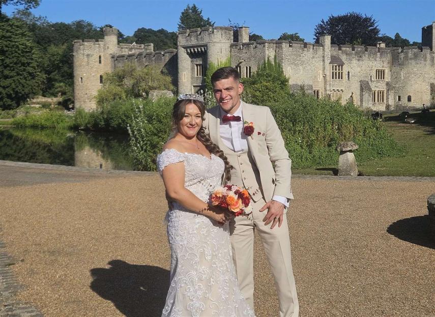 A married couple in wedding attire stand in front of a stone castle.