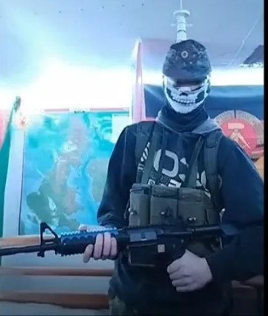 A person with a skull mask and military-style vest holding a rifle.