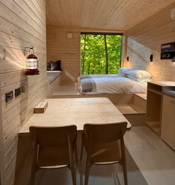 Interior of a small cabin with light wood paneling, featuring a bed, small table with two chairs, and a window looking out onto green trees.