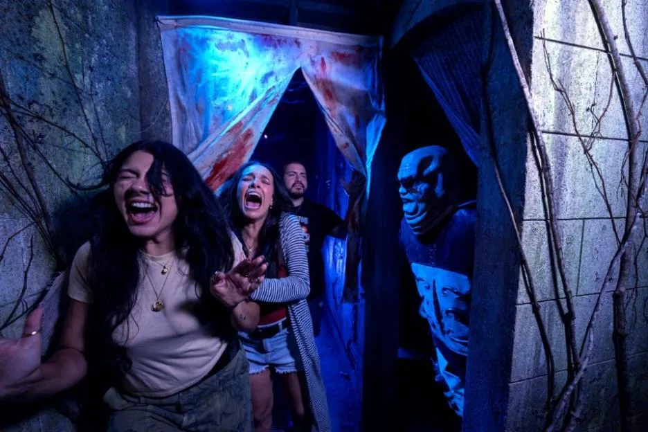 Three people screaming in terror as an alien-like figure approaches them in a haunted house.