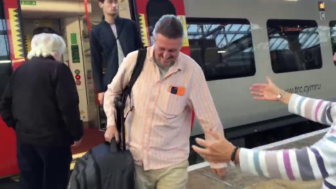 Man smiling and walking towards a woman after stepping off a train.