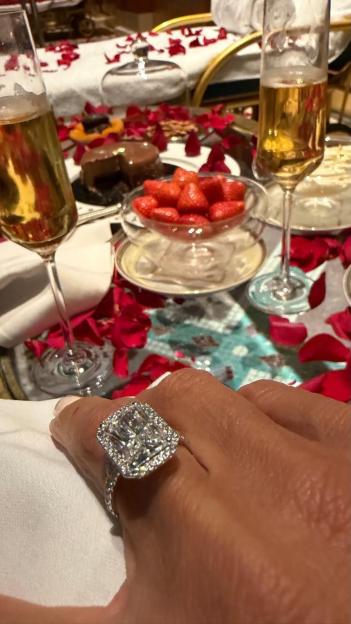 A woman in a white robe stands beside a pool decorated with rose petals and candles, forming the words "I Love You."