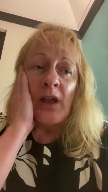 A woman with blonde hair, wearing a black top with white floral patterns, holds her hand to her cheek with an open mouth and distressed expression.