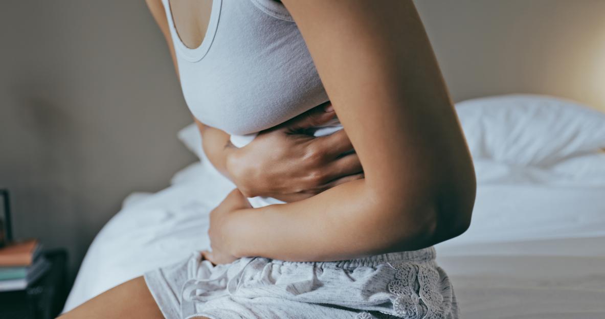 A woman in a white tank top and patterned shorts sits on a bed, clutching her stomach in pain.