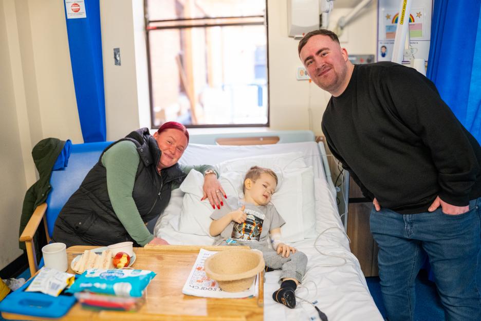 Luke Littler, a woman, and a child in a hospital bed, with Littler leaning in from the right of the frame.
