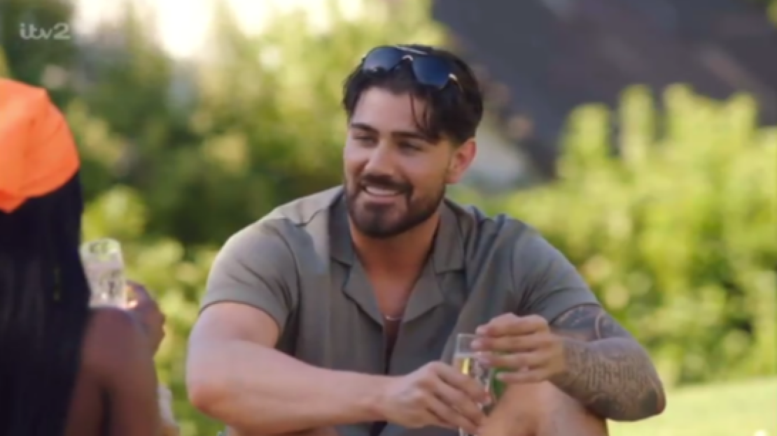 A man with sunglasses on his head smiling while holding a drink, with the back of another person's head visible.