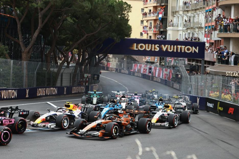Start of a Formula 1 race with numerous cars on the track, surrounded by spectators and buildings.