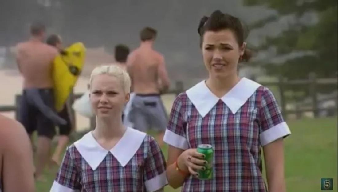 Demi Harman and another actress in plaid school uniforms, with other people on a beach in the background.