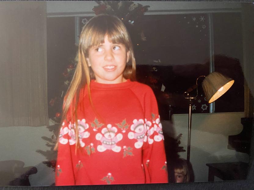 A young girl in a red sweater with teddy bears and holly, looking to the side, with a Christmas tree and window decorated with snowflakes behind her.