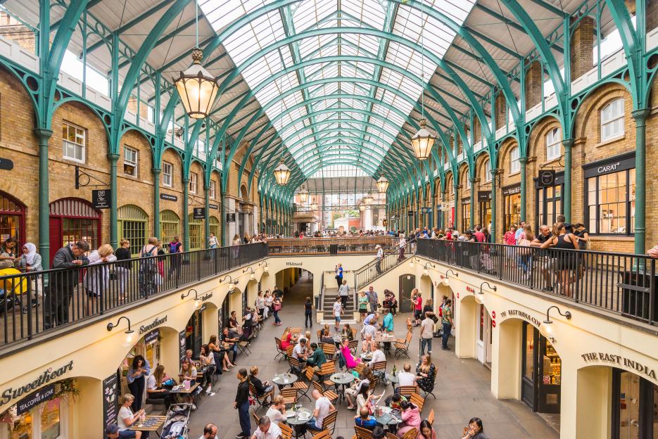 Covent Garden, a two-story flea market in London, filled with people shopping and dining at cafes and restaurants under a glass-arched roof.