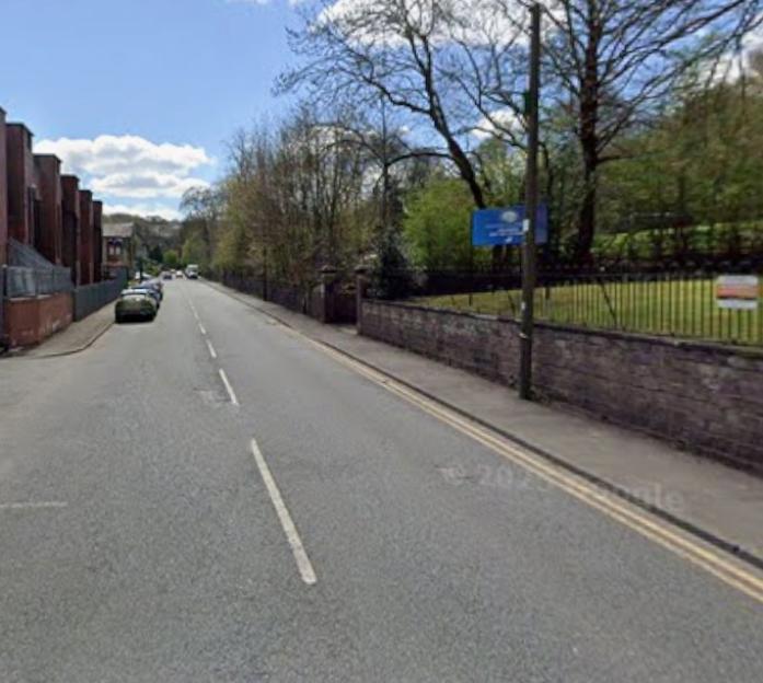 Meltham Mills Road in West Yorkshire, with buildings on the left and trees and a stone wall on the right.