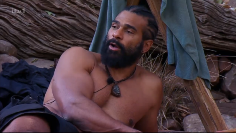 A man with a beard and dark braided hair, wearing a necklace with a pendant, talking in an outdoor setting.