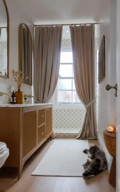 A modern bathroom with a wooden vanity, a large window covered by beige curtains, and a cat lying on the floor.