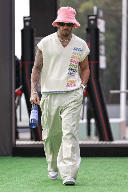 Lewis Hamilton arriving at the track for the F1 Grand Prix of Mexico, wearing a pink bucket hat, sunglasses, a white sweater vest with "DIOR" written repeatedly in rainbow colors, light-colored pants, and holding a blue bottle.