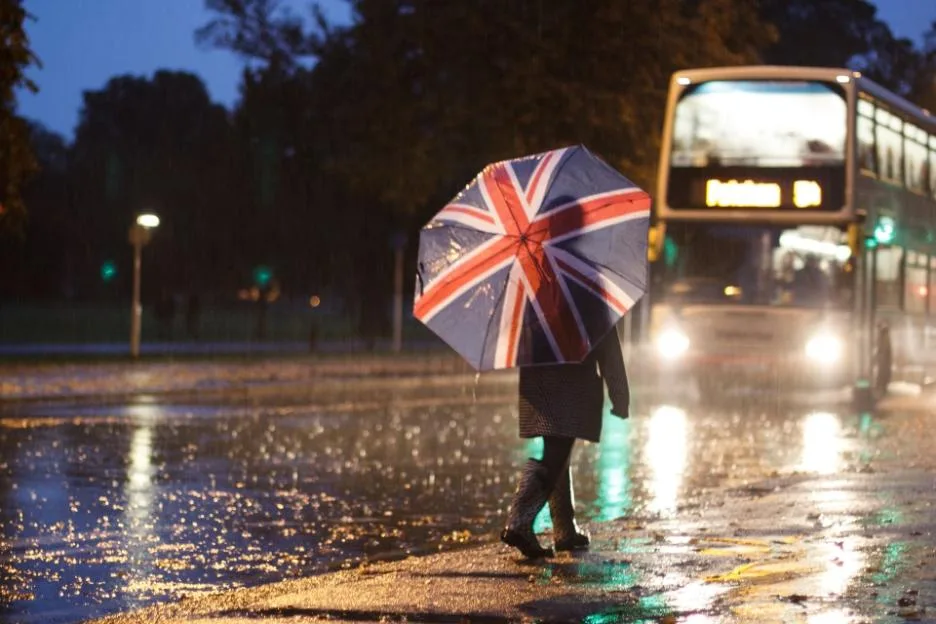 A person walks with a Union Jack umbrella alongside a road on a rainy night as a bus passes by.