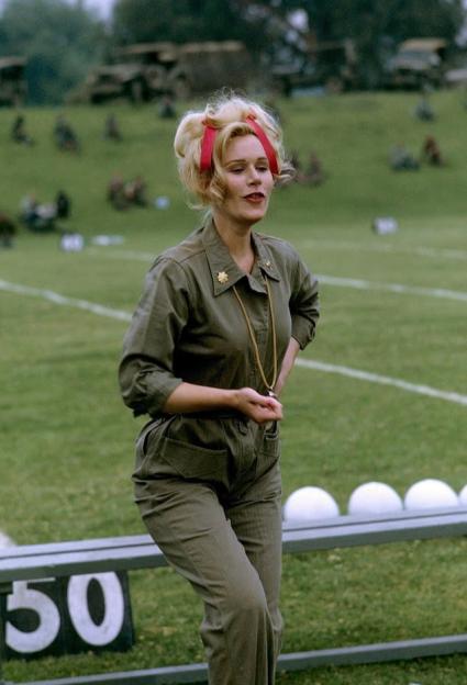 A woman with blonde hair, wearing a green jumpsuit and holding a whistle, stands on a football field.