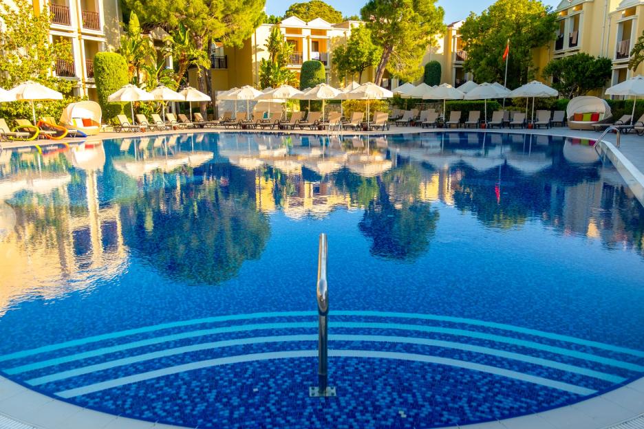 A swimming pool with blue tiles and steps, surrounded by lounge chairs and umbrellas, with a resort building and trees in the background.