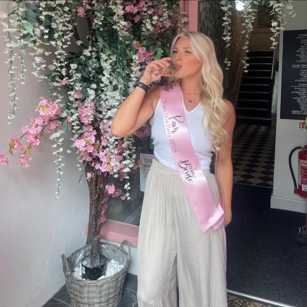Pamela Pritchard, a blonde woman wearing a white tank top, light pants, and a pink "Nia's Hen Party 2025 Team Bride" sash, drinks from a glass while standing in front of pink and white flowers.