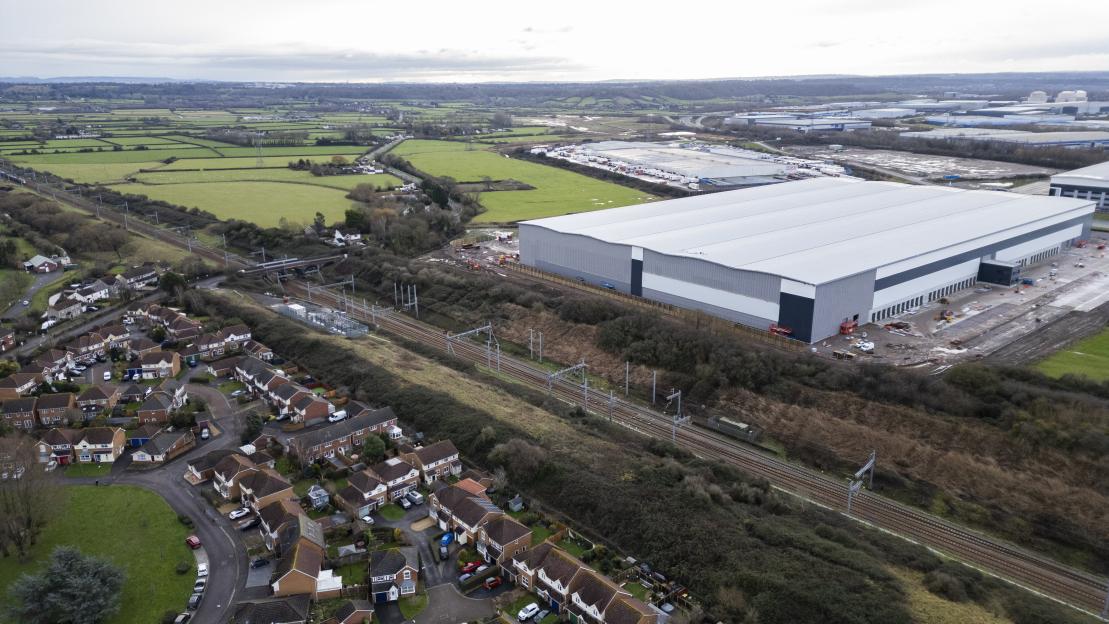 Aerial view of a large, newly constructed warehouse next to a residential area, separated by railroad tracks and fields.