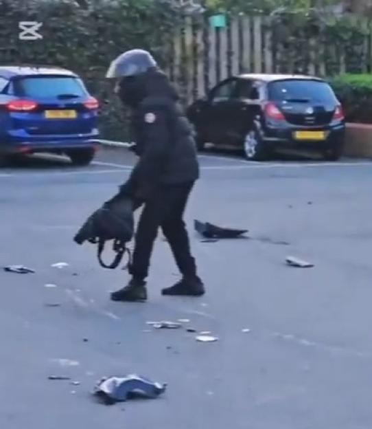 A person in a helmet and dark clothing standing in a car park with debris scattered on the ground.