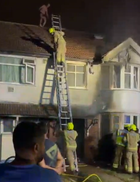 A man on a roof is being helped by a firefighter on a ladder while others stand below.